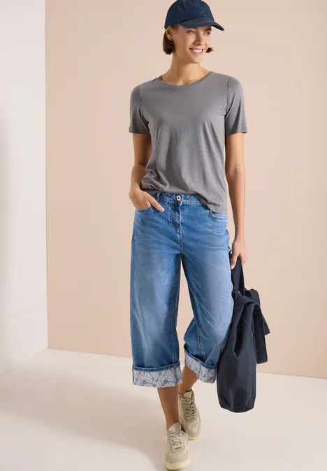 7/8 Wide Legs jeans met omgeslagen detail mid blue wash Vrouw in grijs T-shirt, blauw omslagbroek, sneakers en pet, houdt een spijkerjasje vast voor een lichte achtergrond.