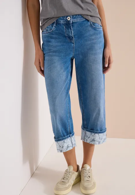 7/8 Wide Legs jeans met omgeslagen detail mid blue wash Persoon draagt blauwe enkellange omgeslagen jeans en beige sneakers voor een lichte muur.