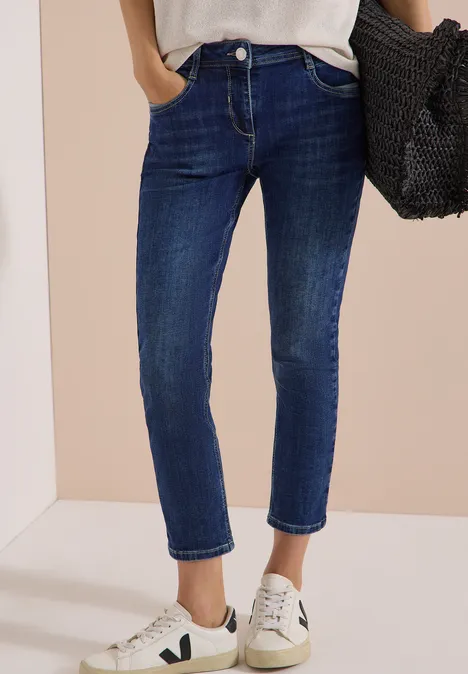 Mid Waist Slim Leg Jeans im Casual Fit mid blue used wash Person trägt dunkelblaue Jeans, weiße Sneakers und hält eine schwarze Tasche in der Hand.