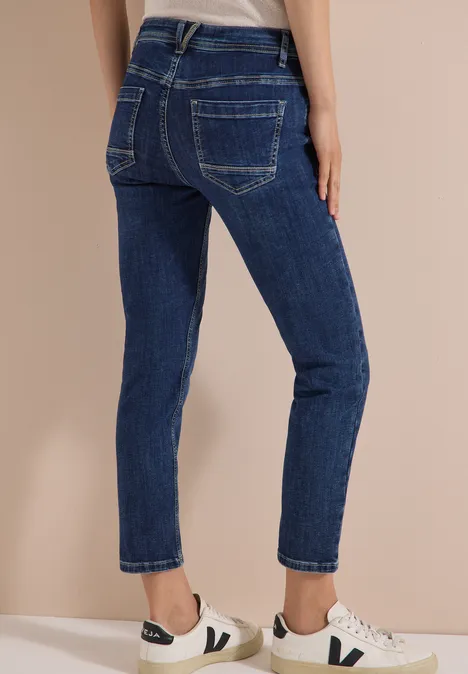 Mid Waist Slim Leg Jeans im Casual Fit mid blue used wash Frau trägt dunkelblaue Jeans und weiße Sneaker, rückseitige Ansicht, beiger Hintergrund.