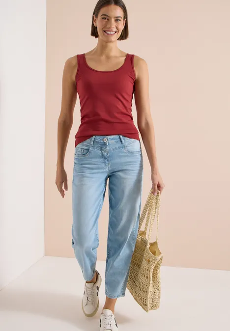 Frau trägt rotes Top, helle Jeans, weiße Sneaker und hält eine geflochtene Strandtasche.
