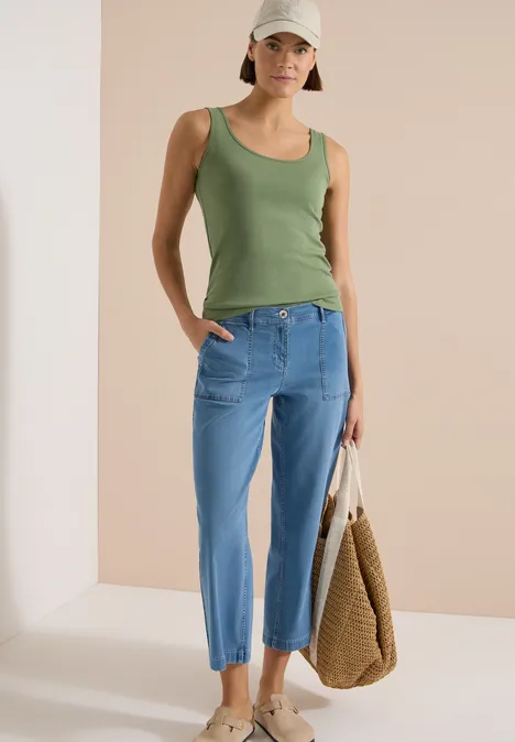 Vrouw in groen topje en losse blauwe jeans, met gevlochten tas voor een neutrale achtergrond.