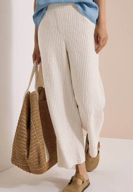 Wide Leg Hose in Seersucker-Qualität grain beige Frau in cremefarbenen, gerippten Hosen mit braunen Sandalen hält eine große geflochtene Tasche.