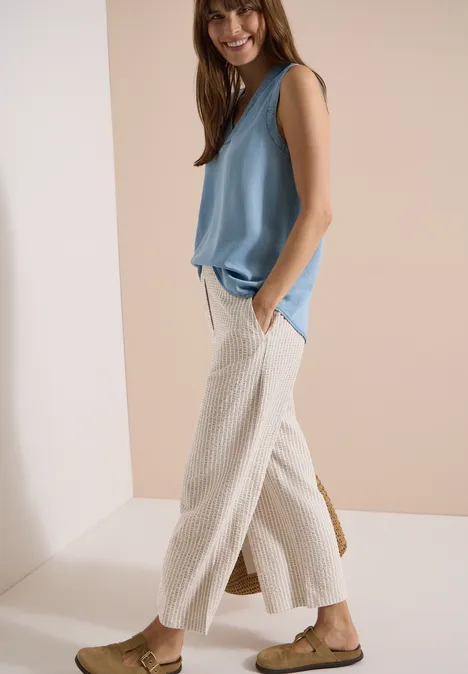 Wide Leg Hose in Seersucker-Qualität grain beige Modell mit ärmelloser hellblauer Bluse, cremeweißer weiter Leinenhose und flachen Sandalen.