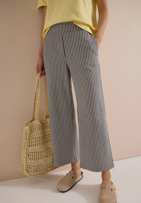 Frau trägt graue, gestreifte Culotte-Hose, gelbes Shirt, Basttasche und beigen Loafer.
