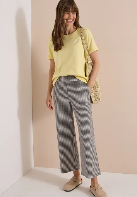 Frau in gelbem T-Shirt, grauen Culottes, flachen Schuhen und gehäkelter Umhängetasche.