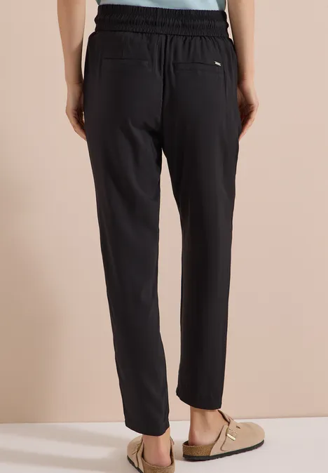 Achteraanzicht van een persoon in losse zwarte broek met elastische taille en zakken, met lichte pantoffels.