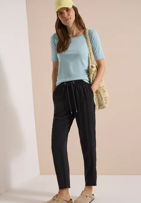 Vrouw in lichtblauw T-shirt en zwarte broek met koord, met strooien tas en sandalen op een neutrale achtergrond.