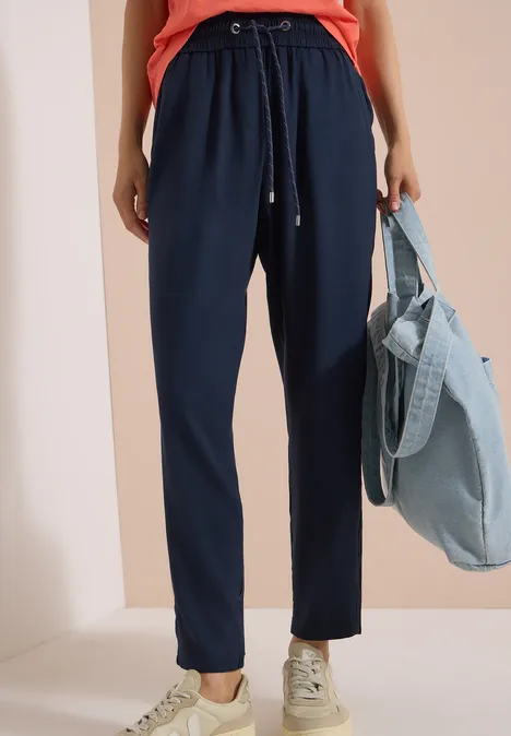 Pantalon Mid Waist Slim Leg en coupe loose universal blue Femme en pantalon bleu marine à cordon, baskets blanches, tenant un sac en jean bleu clair.