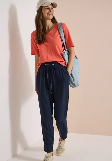 Pantalon Mid Waist Slim Leg en coupe loose universal blue Femme en t-shirt rouge, pantalon de jogging bleu marine et baskets beiges, avec un sac en jean.