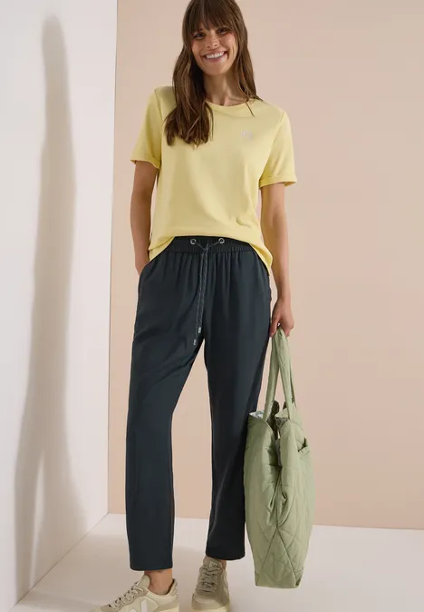 Femme en t-shirt jaune, pantalon sombre, portant un sac vert devant un mur beige.