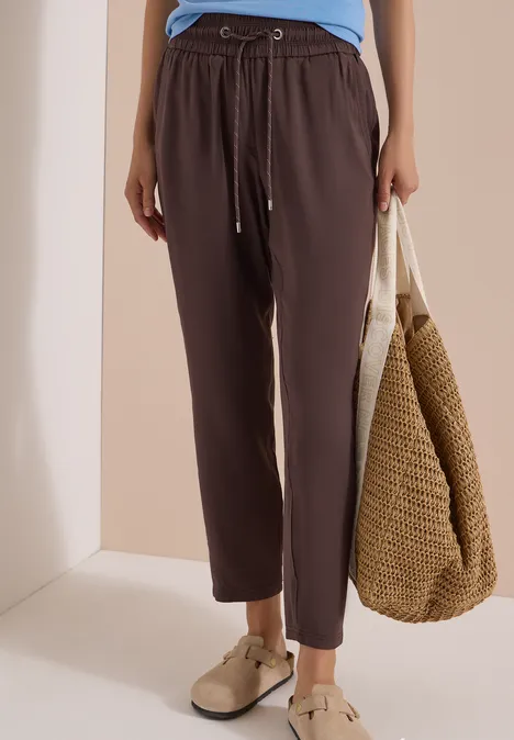Donkerbruine linnenbroek met tunnelkoord, nonchalant model, gecombineerd met strooien tas en beige espadrilles.