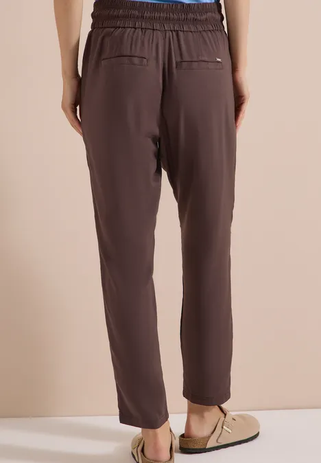 Donkerbruine, hoog getailleerde broek met elastische tailleband en taps toelopende pijpen, gecombineerd met loafers.
