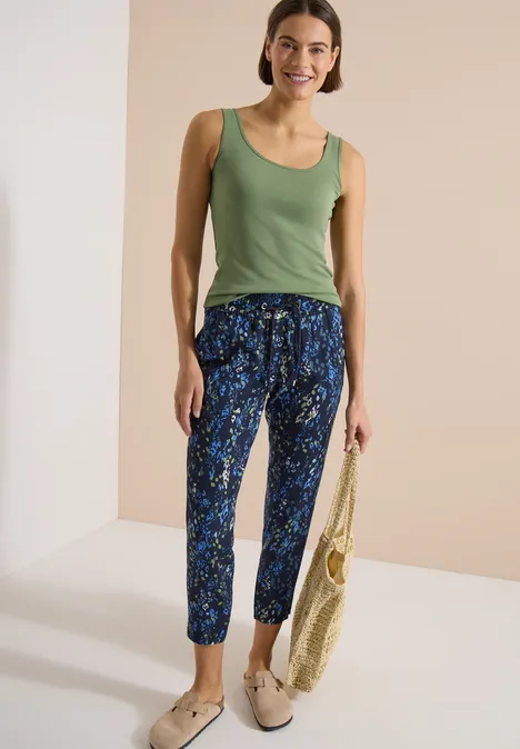 Une femme porte un débardeur vert, un pantalon bleu à motifs et des mocassins beiges, tenant un sac.