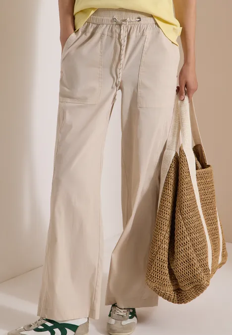 Onderlichaam met wijde lichtbeige broek met koord, groene sneakers en grote gevlochten tas