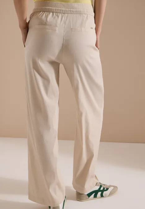 Rugaanzicht van persoon in beige, wijde broek met elastische taille, handen in zakken, witte sneakers met groene strepen.