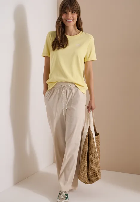 Vrouw in een los geel T-shirt en beige linnenbroek met een gevlochten tas, staand voor een neutrale achtergrond.