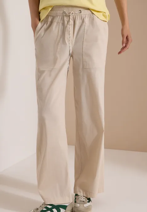 Onder aanzicht van iemand in wijde beige broek met elastische taille, koord en witte sneakers.
