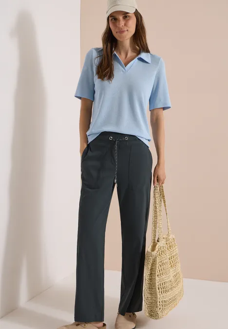 Vrouw draagt lichtblauw poloshirt, wijde donkere broek en houdt een stro tas vast.