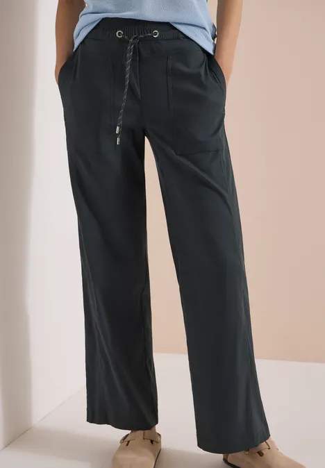 Losvallende donkergrijze broek met trekkoord en zakken, gecombineerd met bruine schoenen.