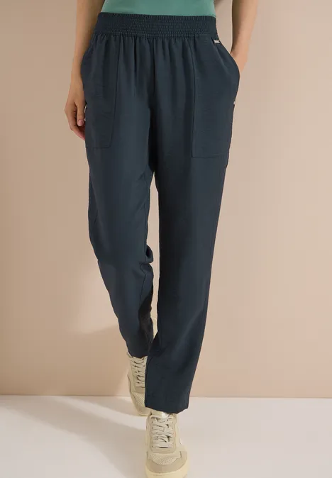 Lockere dunkelblaue Hose mit großen Taschen und weißen Sneakern, getragen vor schlichtem beigem Hintergrund.