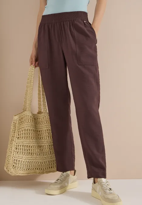 Frau trägt braune, lässige Hose, cremefarbenes Top, beige Sneaker und gehäkelte Tasche.