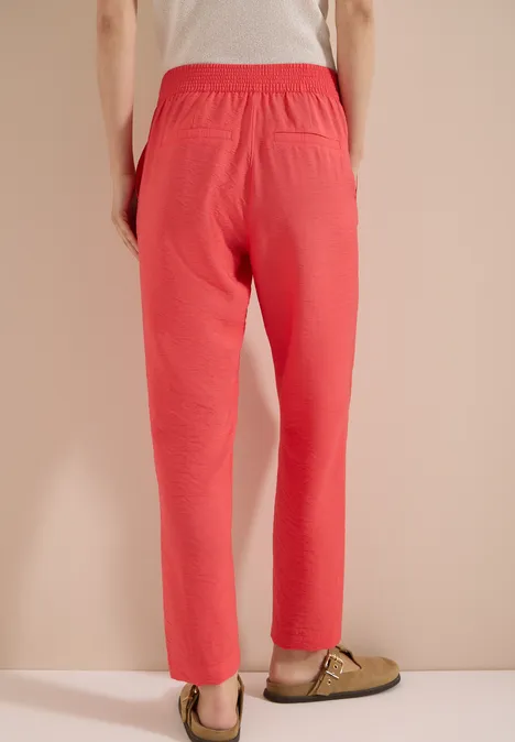 Person steht vor beiger Wand und trägt eine korallrote Hose mit grünen Pantoffeln.