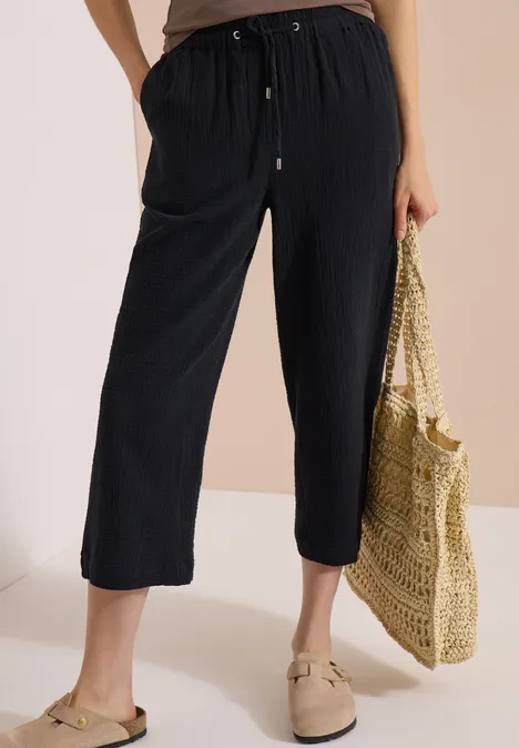 3/4 Wide Leg Hose in Musselin-Qualität Black Frau in schwarzen, knöchellangen Hosen, beigen Slippern und einer geflochtenen Strohtasche.