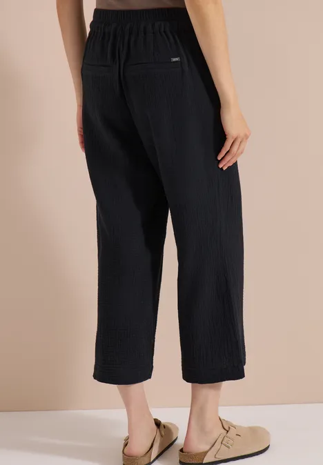 3/4 Wide Leg Hose in Musselin-Qualität Black Rückenansicht: schwarze, weite, knöchellange Rippstrick-Hose mit elastischem Bund und hellen Slippers.