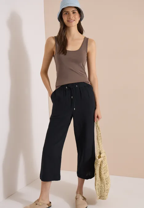 3/4 Wide Leg Hose in Musselin-Qualität Black Frau in schwarzer 3/4-Hose, braunem Top, Strohtasche und Bucket Hat vor schlichtem Hintergrund.