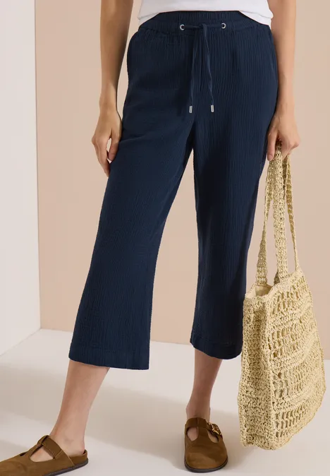 Pantalon 7/8 bleu foncé, ample et texturé, associé à des sandales marron et un sac filet en paille.