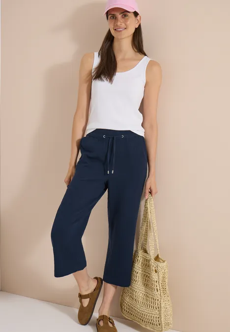 Une femme porte un top blanc, des culottes bleu marine, des sandales et tient un sac en paille.