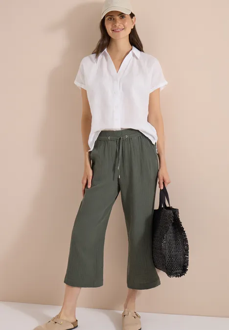 3/4 Wide Leg Hose in Musselin-Qualität clear khaki Frau in weißer Kurzarmbluse, olivgrünen Culotte-Hosen und schwarzer Flecht-Tasche vor beigem Hintergrund.