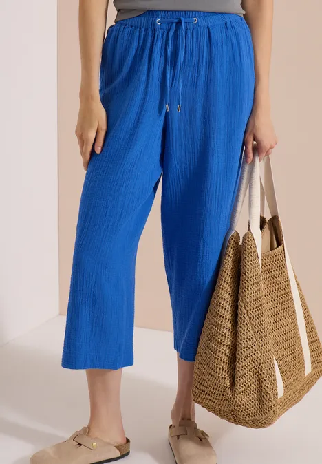 3/4 Wide Leg Hose in Musselin-Qualität surf blue Eine Person trägt knöchellange, weite, blaue Hose mit Kordelzug, flache Sandalen und hält eine Strohtasche.