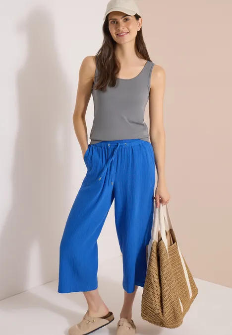 3/4 Wide Leg Hose in Musselin-Qualität surf blue Frau in grauem Tanktop und blauer Culotte, Sandalen und geflochtener Tasche vor hellem Hintergrund.
