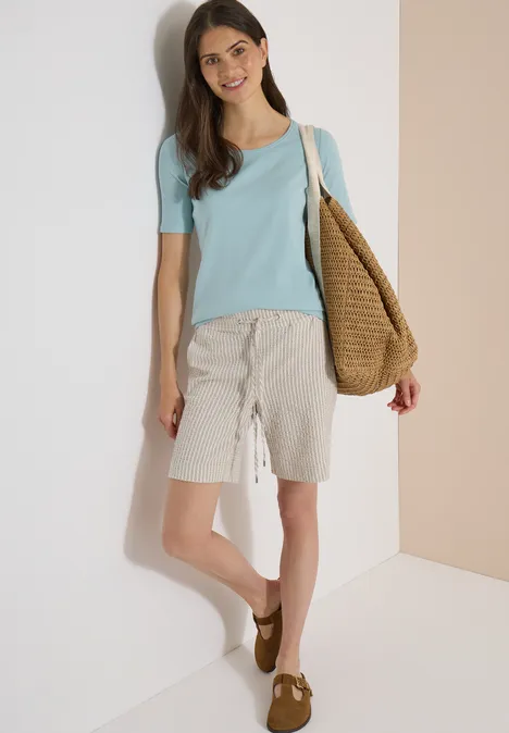 Eine Frau trägt ein hellblaues T-Shirt, beige Shorts, braune Schuhe und eine Strohtasche und lehnt an einer Wand.