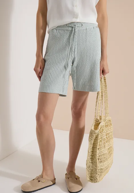 Frau in weißer Bluse, hellblauen Streifen-Shorts, Espadrilles und Strohtasche.