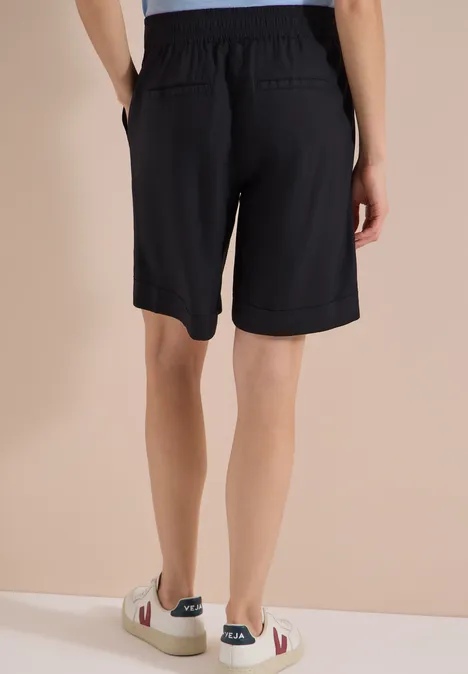Rückansicht einer Person in schwarzen Bermudashorts und weißen Sneakern vor neutralem Hintergrund.
