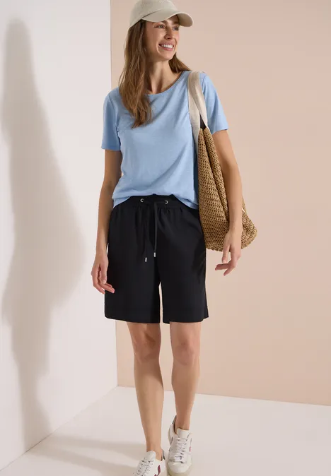 Frau in blauem T-Shirt, schwarzen Bermuda-Shorts, weißen Sneakers, Cap und Basttasche vor neutralem Hintergrund.