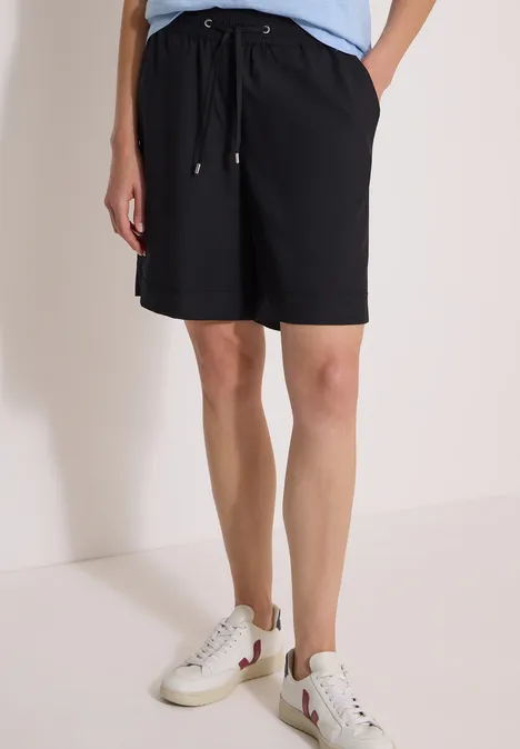 Model trägt schwarze knielange Shorts mit Tunnelzug und weiße Sneaker.