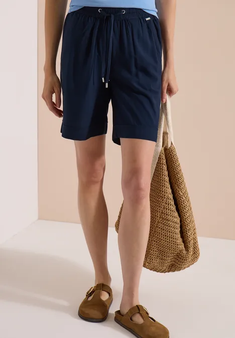 High Waist Wide Leg Shorts im Loose Fit universal blue Frau trägt marineblaue Shorts mit Kordelzug und braunen Sandalen, hält eine geflochtene Tasche.