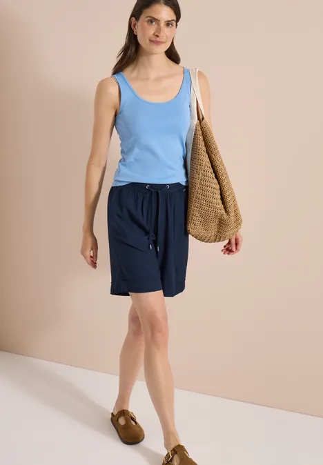 High Waist Wide Leg Shorts im Loose Fit universal blue Frau in blauem Tanktop, dunkelblauen Shorts und braunen Sandalen vor beigem Hintergrund mit großer Gewebetasche.