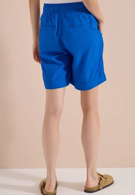 High Waist Wide Leg Shorts im Loose Fit surf blue Eine Person von hinten in knielangen, leuchtend blauen Shorts und hellen Slippern vor einem neutralen Hintergrund.