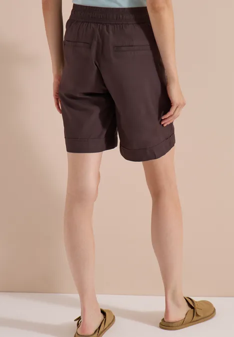 High Waist Wide Leg Shorts im Loose Fit brown cacao Von hinten gezeigte Person in dunkelbraunen Shorts und grünen Slippern vor beigem Hintergrund.
