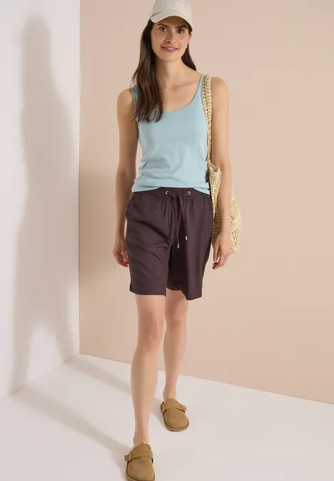 High Waist Wide Leg Shorts im Loose Fit brown cacao Frau in türkisfarbenem Tanktop, braunen Shorts mit Kordelzug und braunen Sandalen vor hellem Hintergrund.