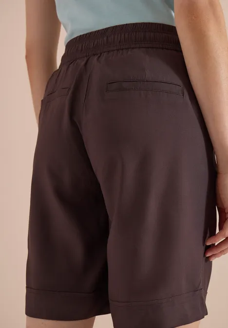 High Waist Wide Leg Shorts im Loose Fit brown cacao Frau von hinten in dunkelbraunen, knielangen Shorts mit Gummibund und Tasche, dazu hellblaues Oberteil.