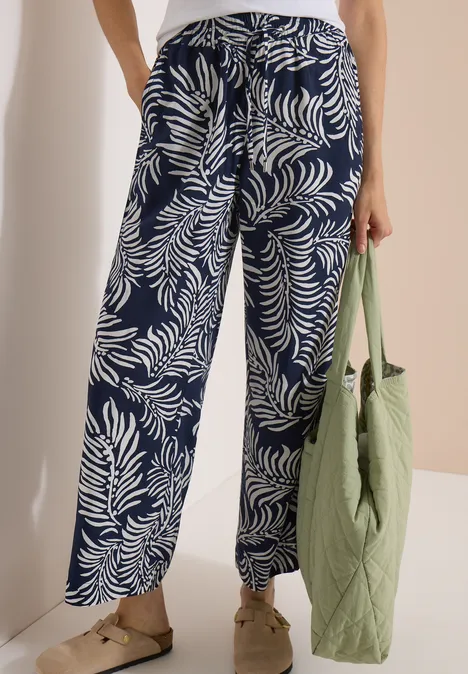 Une personne porte un pantalon large bleu marine à motifs de fougères blanches et tient un sac matelassé vert.