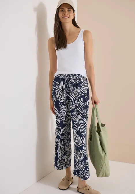 Femme en débardeur blanc et pantalon ample bleu et blanc à motif de palmiers, sac vert et chaussures beiges.