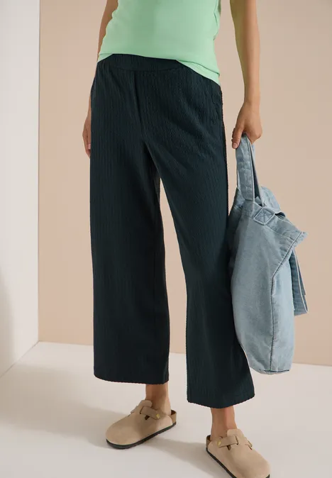 Vrouw in wijd, knielang broek, groene top, met een spijkerjasje in de hand en beige muiltjes.