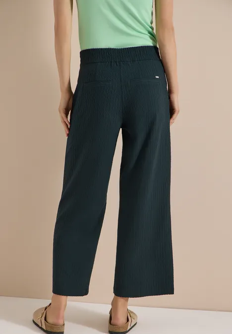 Rugaanzicht van een vrouw in wijd donkeblauwe, enkellange broek, groen T-shirt en platte sandalen.